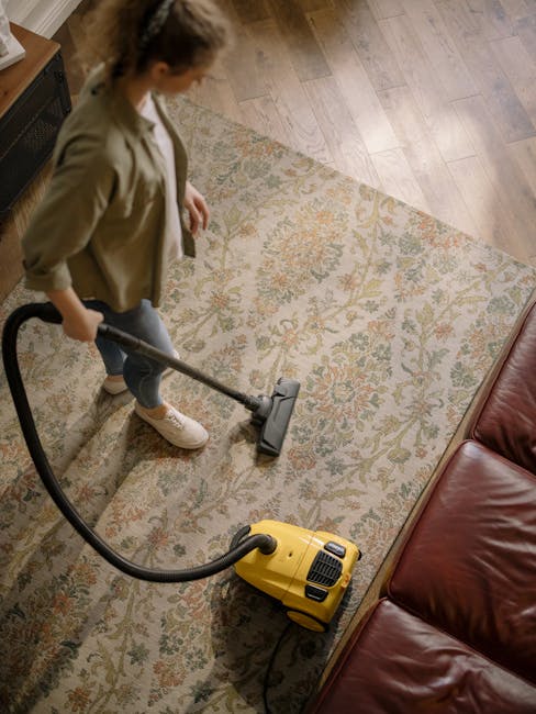A woman operating a yellow vacuum cleaner on a patterned, beige and multicolored carpet in a living room with wooden flooring visible at the edges. The room features a red leather sofa partially visib