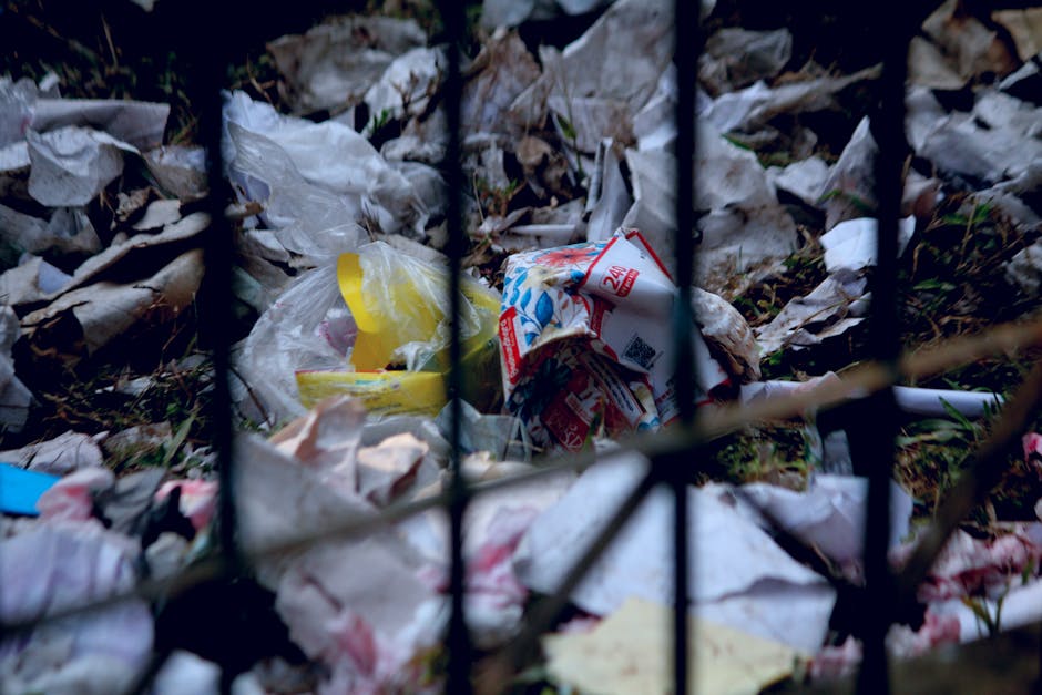 A close-up view of a pile of various household waste, including crumpled paper, plastic bags, and food packaging, scattered on the ground behind a black metal fence. The waste appears to be outside, w