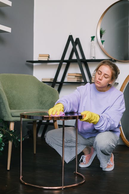 A person wearing black gloves is using a handheld electric upholstery steamer to deep clean a dark grey fabric sofa in a living room. The sofa has a modern design with straight lines and a cushioned b
